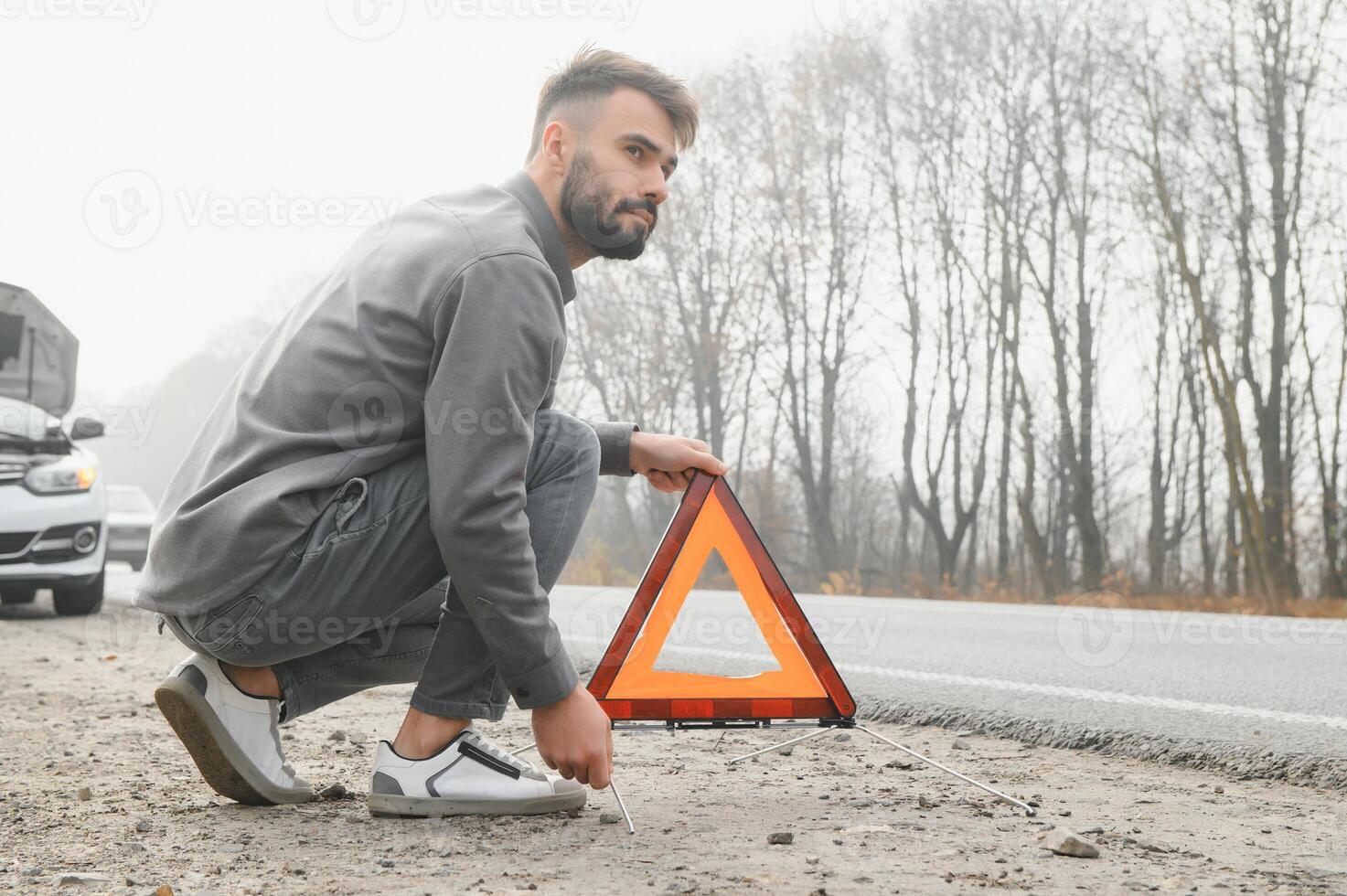 A young man near a broken car with an open hood on the roadside. photo