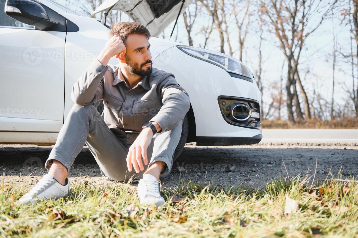 Frustrated sad upset guy driver is sitting near a broken car after road accident, crash. Young shocked desperate frightened man got into traffic car accident, sitting on asphalt holding head with hand photo