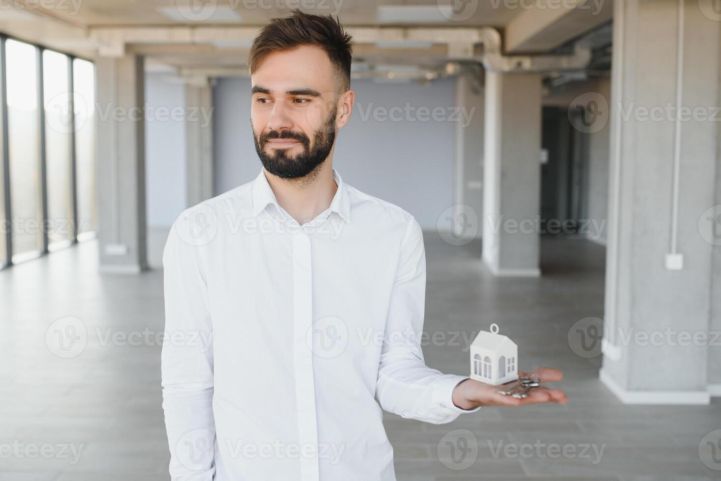 Indoor portrait of happy male home owner showing key to his new house. Joyful handsome single young Caucasian man in casual shirt standing in room, holding keys and smiling. Real estate concept photo