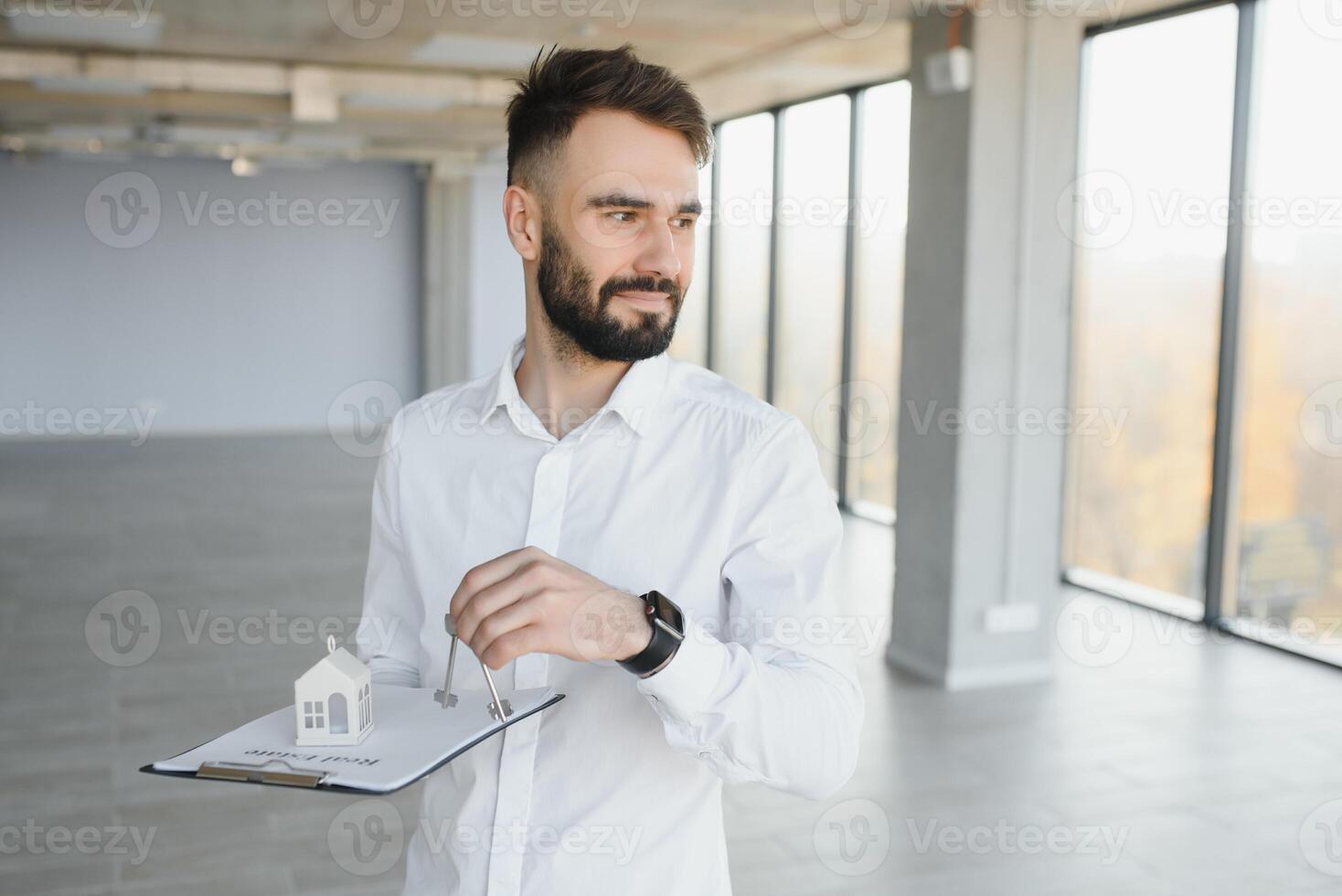 Indoor portrait of happy male home owner showing key to his new house. Joyful handsome single young Caucasian man in casual shirt standing in room, holding keys and smiling. Real estate concept photo
