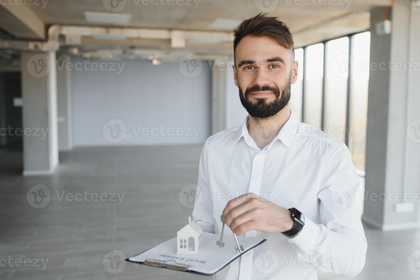 Indoor portrait of happy male home owner showing key to his new house. Joyful handsome single young Caucasian man in casual shirt standing in room, holding keys and smiling. Real estate concept photo