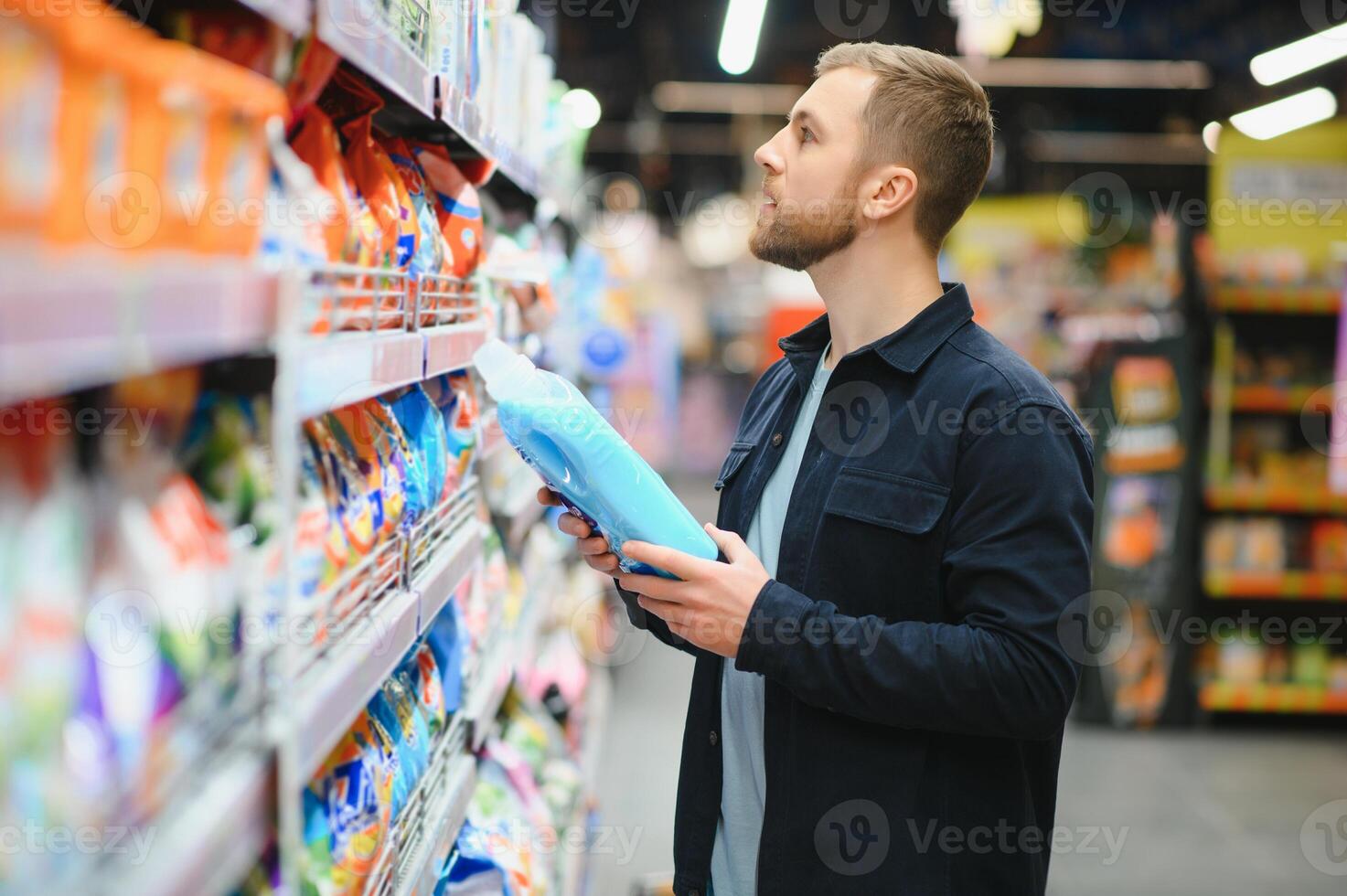 Young man in the supermarket in the household chemicals department. Large selection of products. A brunette in a glasses and a beard in a beige coat. photo