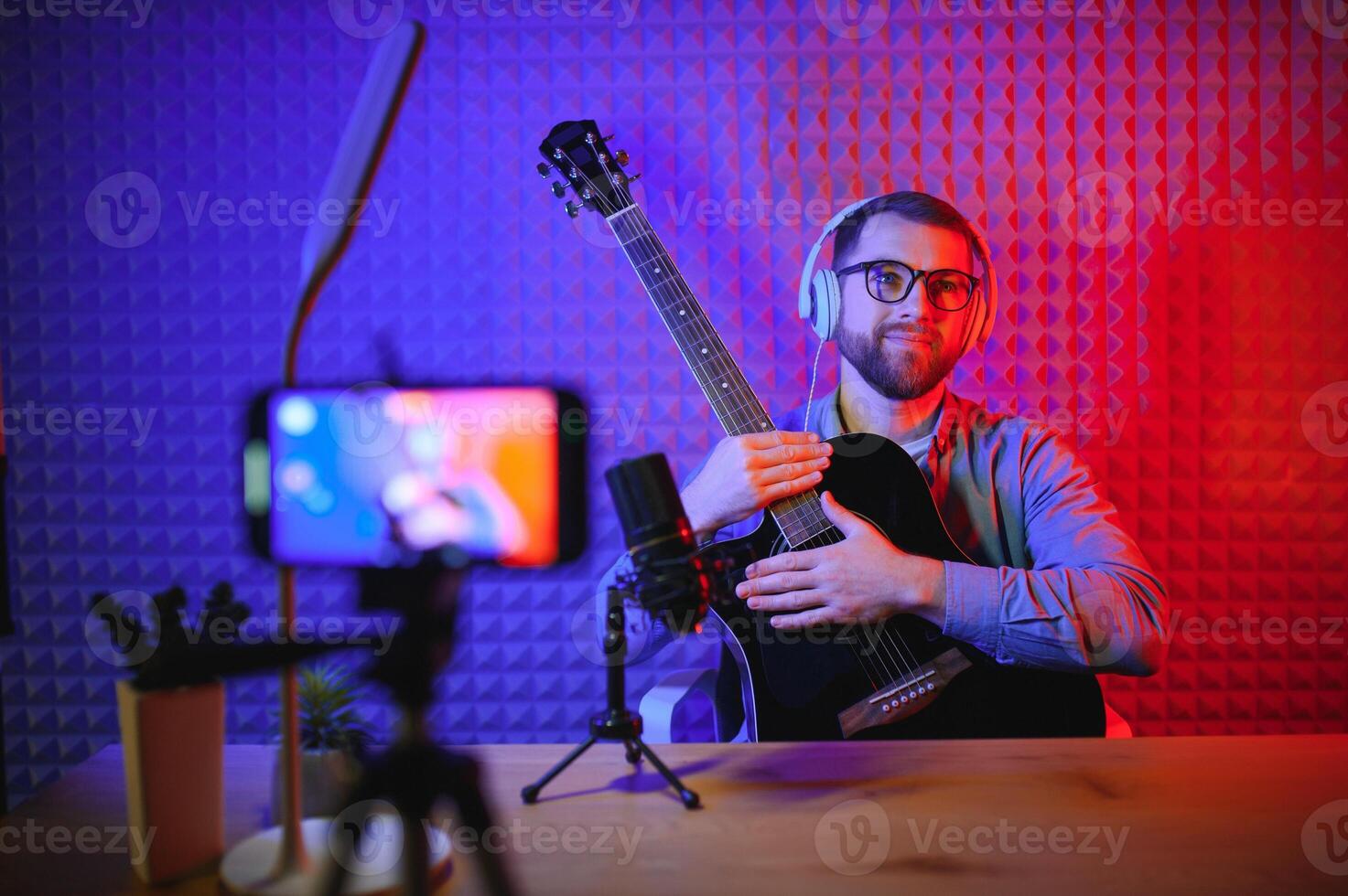Young Caucasian male singer in headphones hold play guitar record new single on smartphone at home studio. Millennial man artist use musical instrument sing shoot music on cellphone camera. photo
