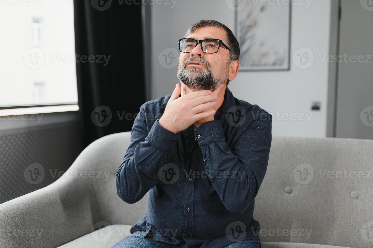 Senior man suffering from cold. Photo of elderly man having sore throat. Elderly Caucasian man touching his throat. Sore throat, tonsillitis or thyroid gland problem. Elder man at home feeling unwell.