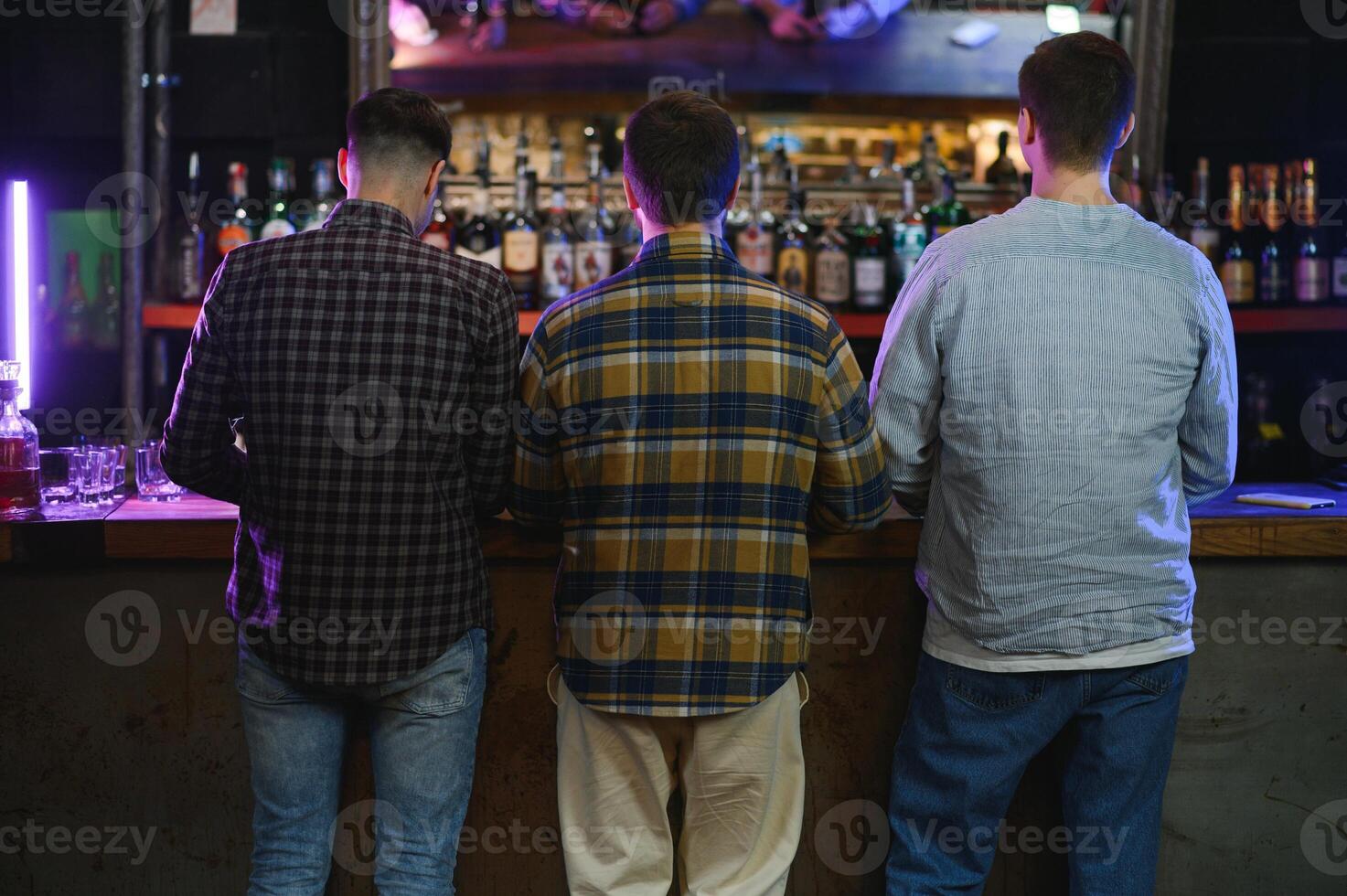 Three young men in casual clothes are smiling, holding bottles of beer while standing near bar counter in pub photo