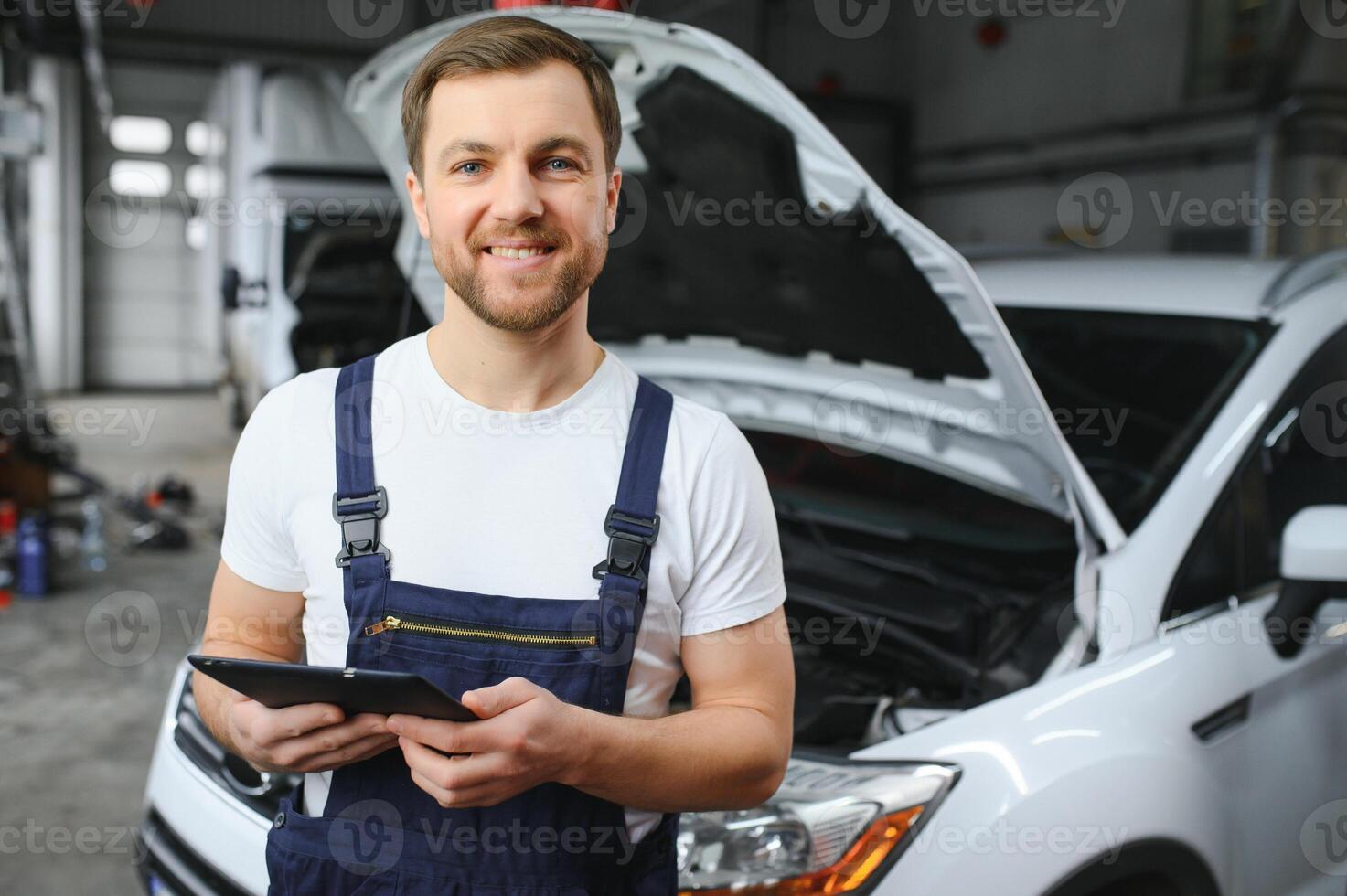 Bearded mechanic in overalls standing in garage of a car salon and holding tablet. He is about to diagnostic breakdown. photo