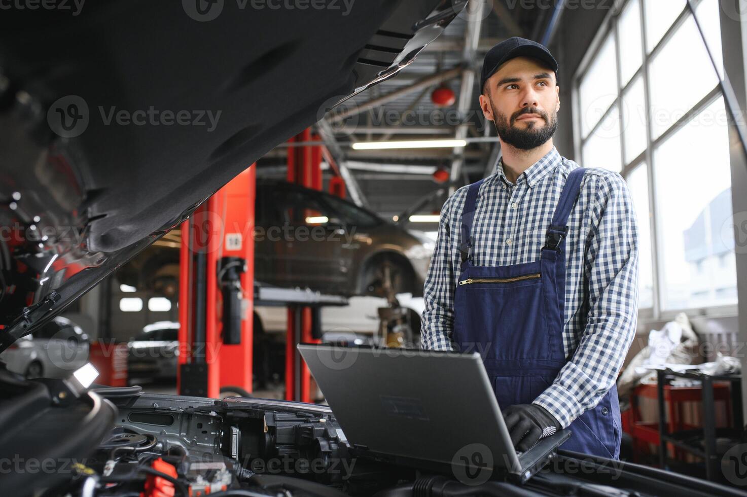 A bearded auto-mechanic in working uniform is standing next to a broken car and using the special laptop for car maintenance. An auto-mechanic in workshop repairs car. photo