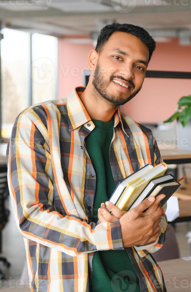 Happy smart indian or arabian guy, mixed race male, university student, stands in the library against the background of bookshelves, holds a lot of books in his hands photo