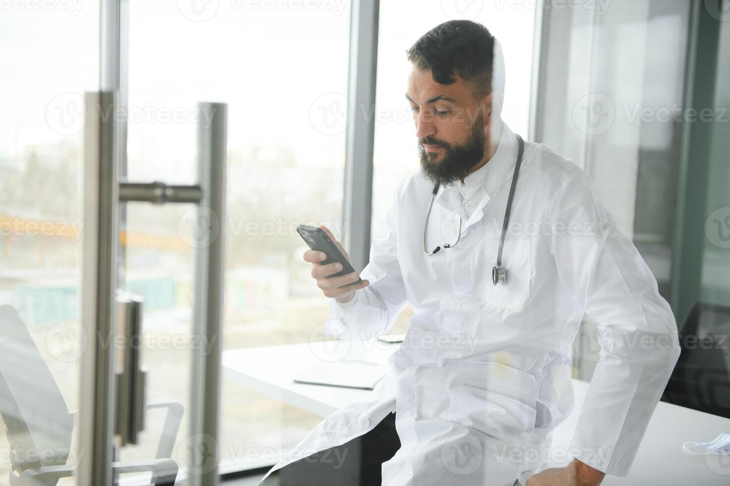 Young 30-aged handsome pleasant Arabic male doctor in white coat, posing at camera indoors. photo