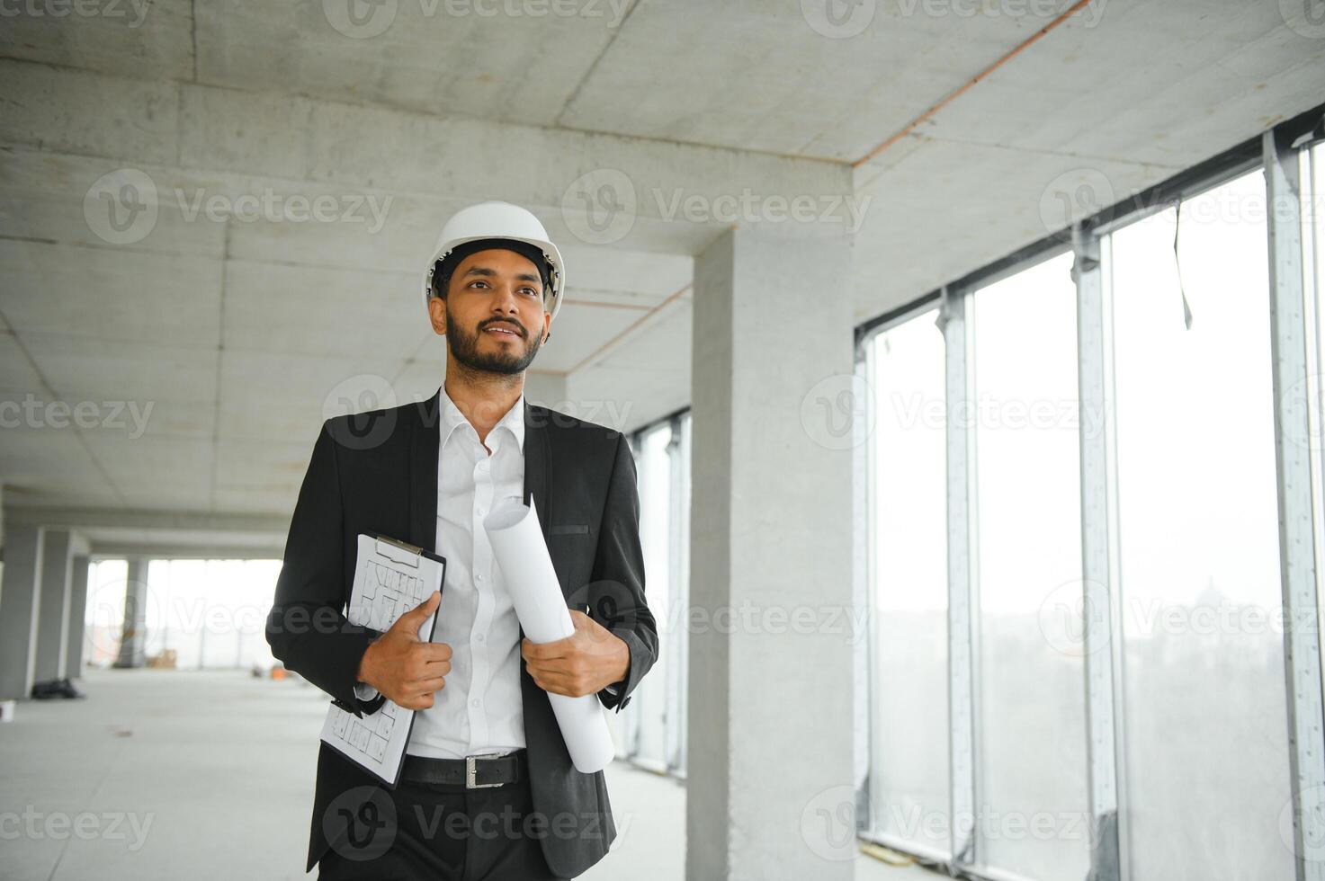 Indian construction site manager standing wearing helmet, thinking at construction site. Portrait of mixed race manual worker or architect. photo