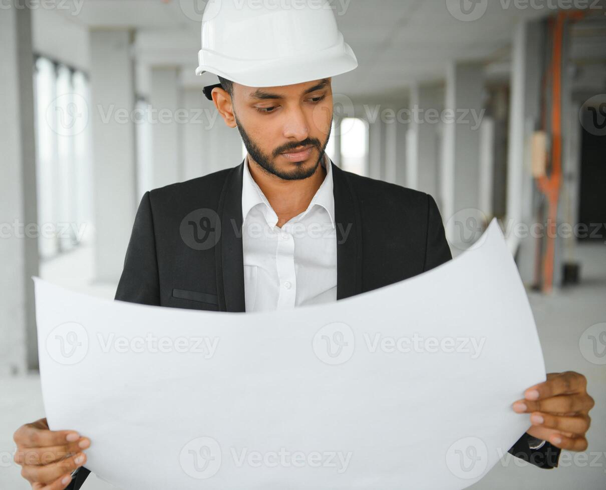 Indian construction site manager standing wearing helmet, thinking at construction site. Portrait of mixed race manual worker or architect. photo
