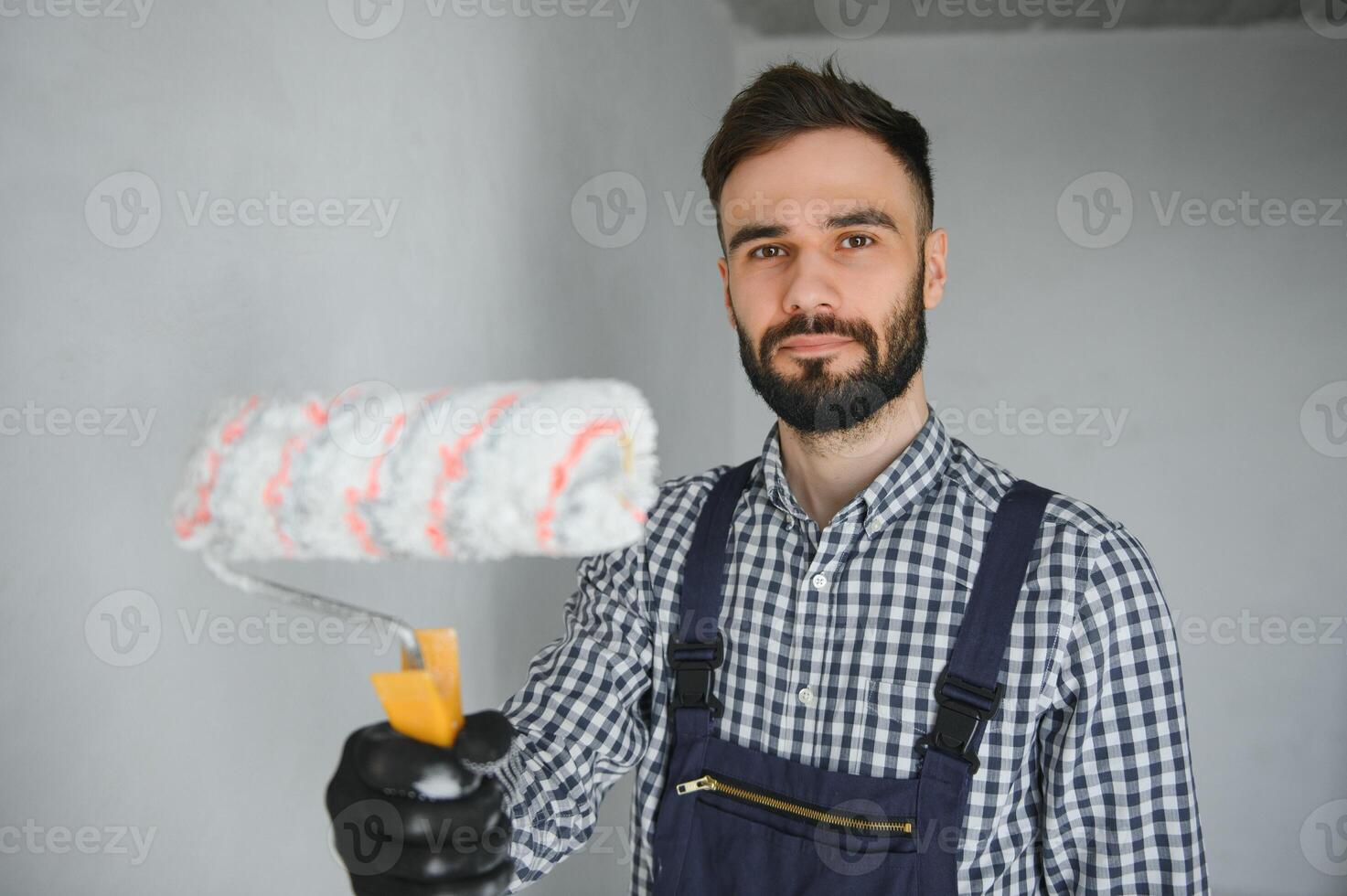 Young smiling professional worker in blue uniform standing with paint roller in new apartment for repairing over grey walls background, copy space. photo