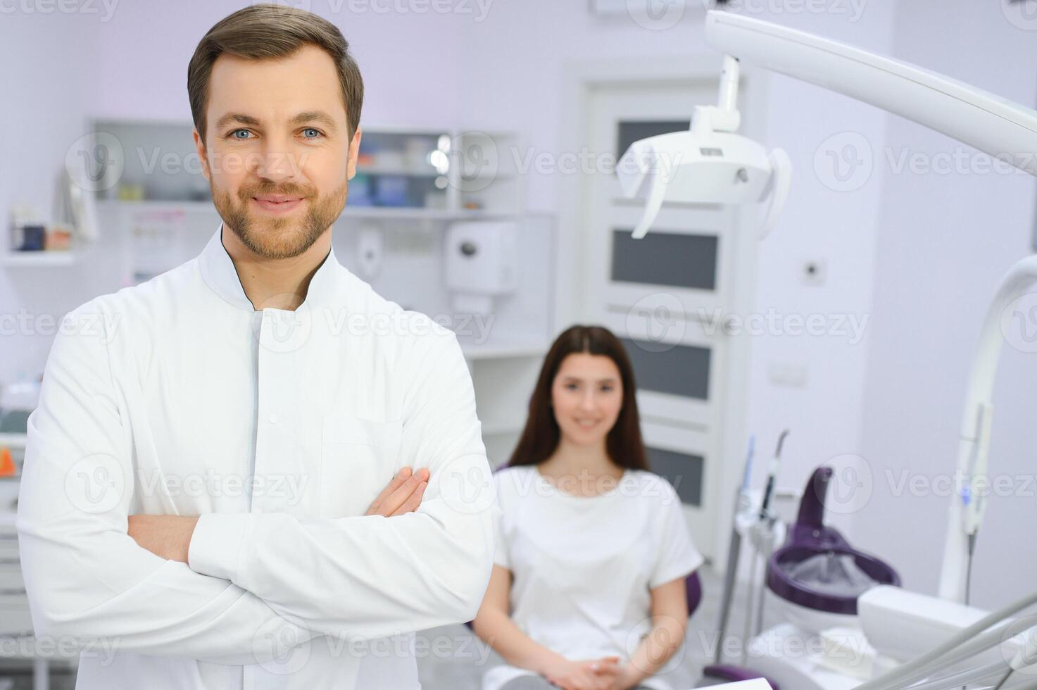 Portrait of positive male dentist with crossed hands posing at camera with patient and nurse on background photo