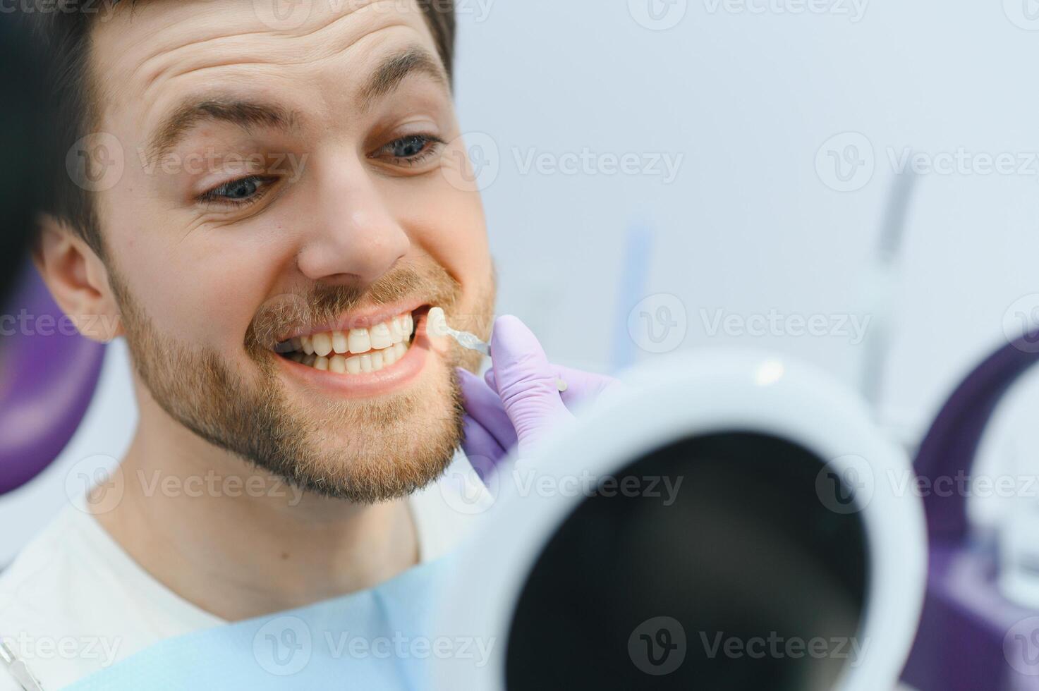 Dentist in blue medical gloves applying sample from tooth enamel scale to smiling man patient teeth to pick up right shade, teeth bleaching procedure photo