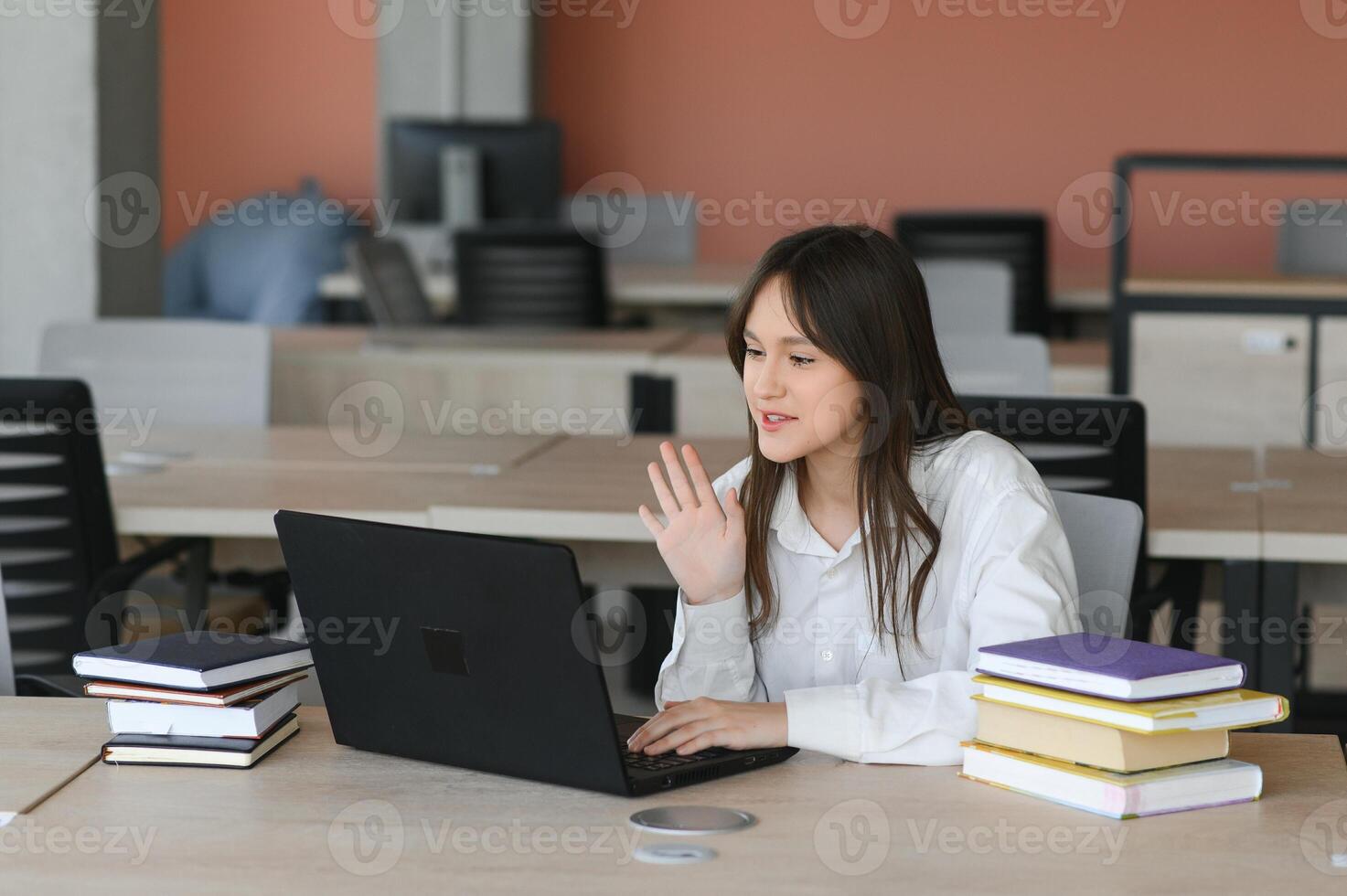 A beautiful caucasian female student is studying in college remotely. She is sitting with a laptop and a notepad and concentrated is watching a conference lesson photo