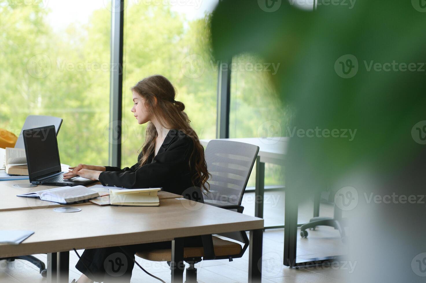A beautiful caucasian female student is studying in college remotely. She is sitting with a laptop and a notepad and concentrated is watching a conference lesson photo