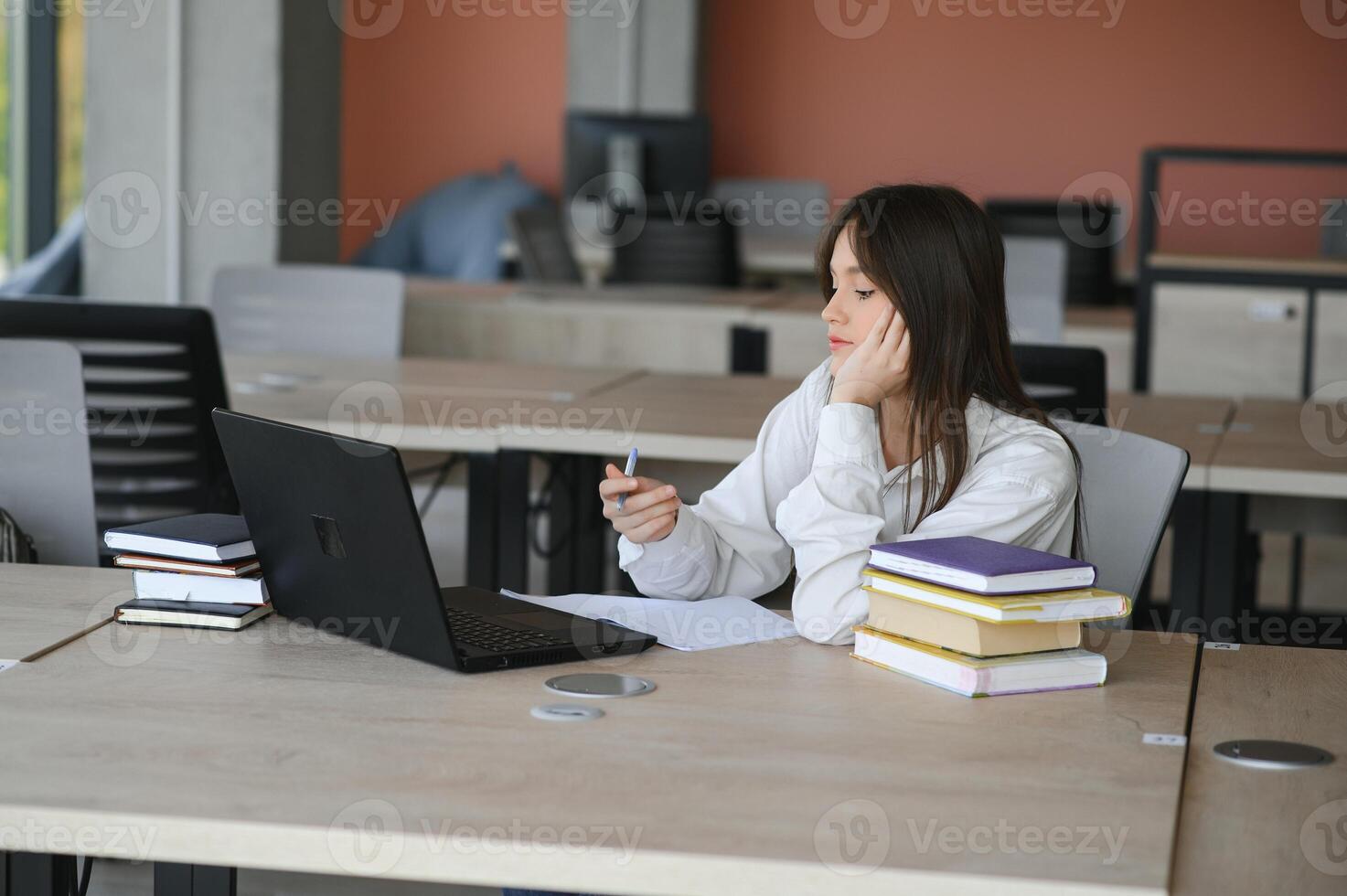 A beautiful caucasian female student is studying in college remotely. She is sitting with a laptop and a notepad and concentrated is watching a conference lesson photo