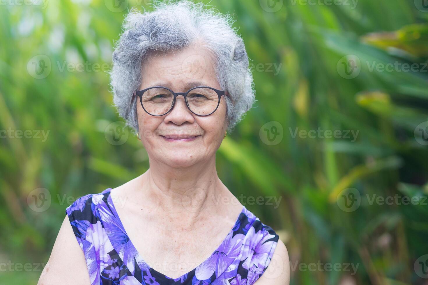 Portrait of a senior woman with short gray hair, wearing glasses, smiling and looking at the camera while standing in a garden. Aged people and relaxation concept photo