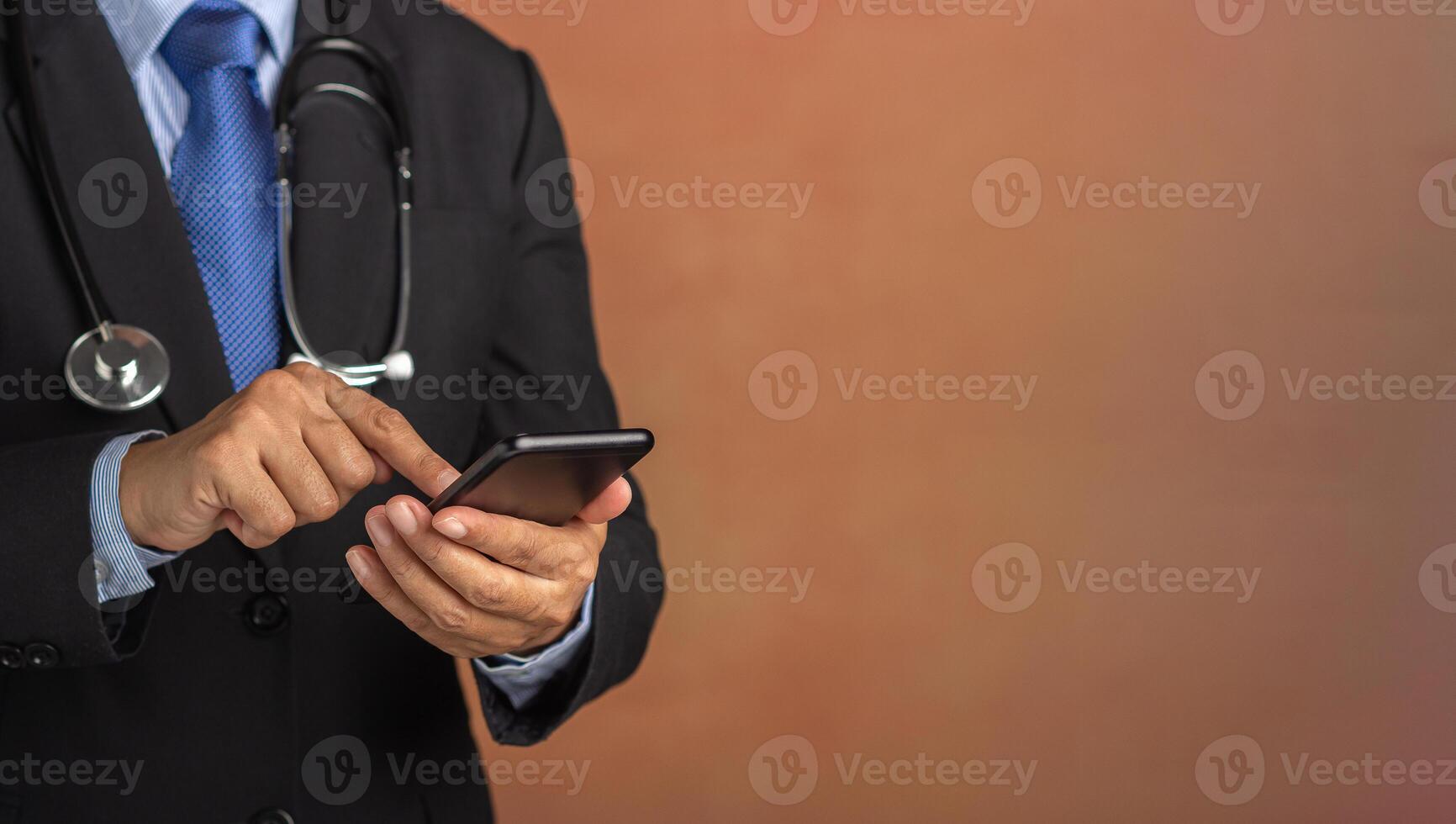 Doctor with stethoscope and dressing in a business suit using smartphone while standing with a brown background. Space for text. Medical and communication concept photo