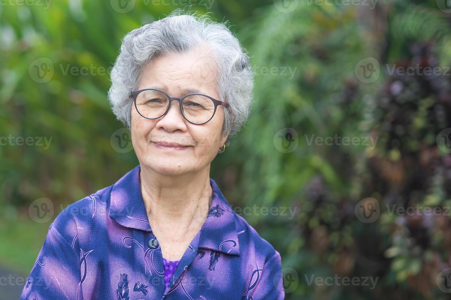 Cheerful grandmother with short gray hair wearing glasses, smiling and looking at the camera while standing in a garden. Space for text. Concept of aged people and healthcare photo