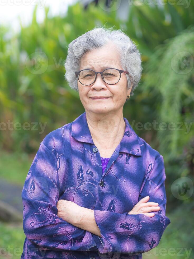 Portrait of a senior woman with short gray hair smiling, arms crossed and looking at the camera standing in a garden. Concept of aged people and relaxation photo