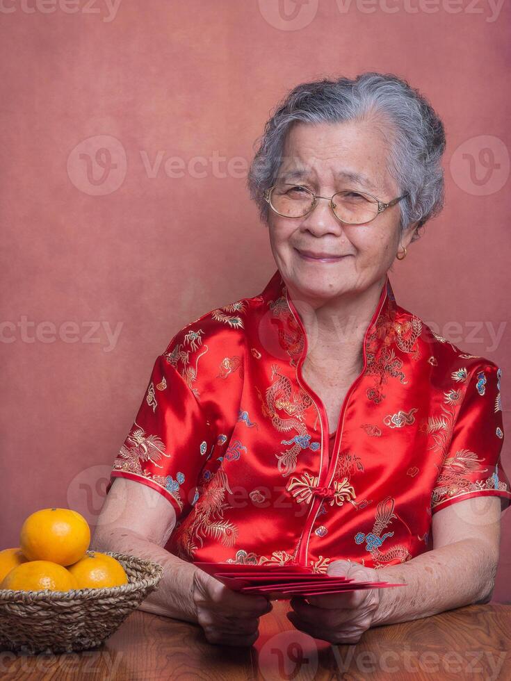 Senior woman wearing a traditional cheongsam qipao dress holding red envelopes and looking at the camera while sitting in the studio with four tangerines in a basket. Happy Chinese New Year photo