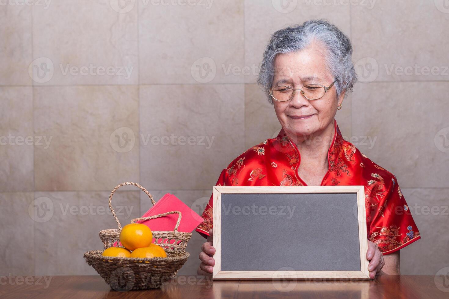 Senior woman wearing a traditional cheongsam qipao dress, smiling, holding, and looking at a blackboard while sitting in the studio with four tangerines and an Ang Pao in a basket. Chinese New Year photo
