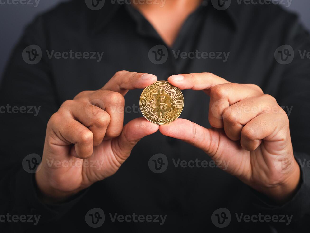 Businessman wearing a black shirt holding a bitcoin coin while standing with a gray background. Close-up photo. Space for text. Virtual cryptocurrency and future money concept photo