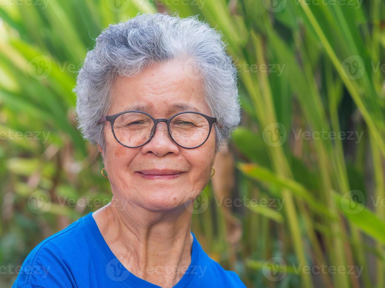 Portrait of a beautiful senior woman with short gray hair wearing glasses, smiling with arms crossed, and looking at the camera while standing in a garden. Concept of aged people and healthcare photo