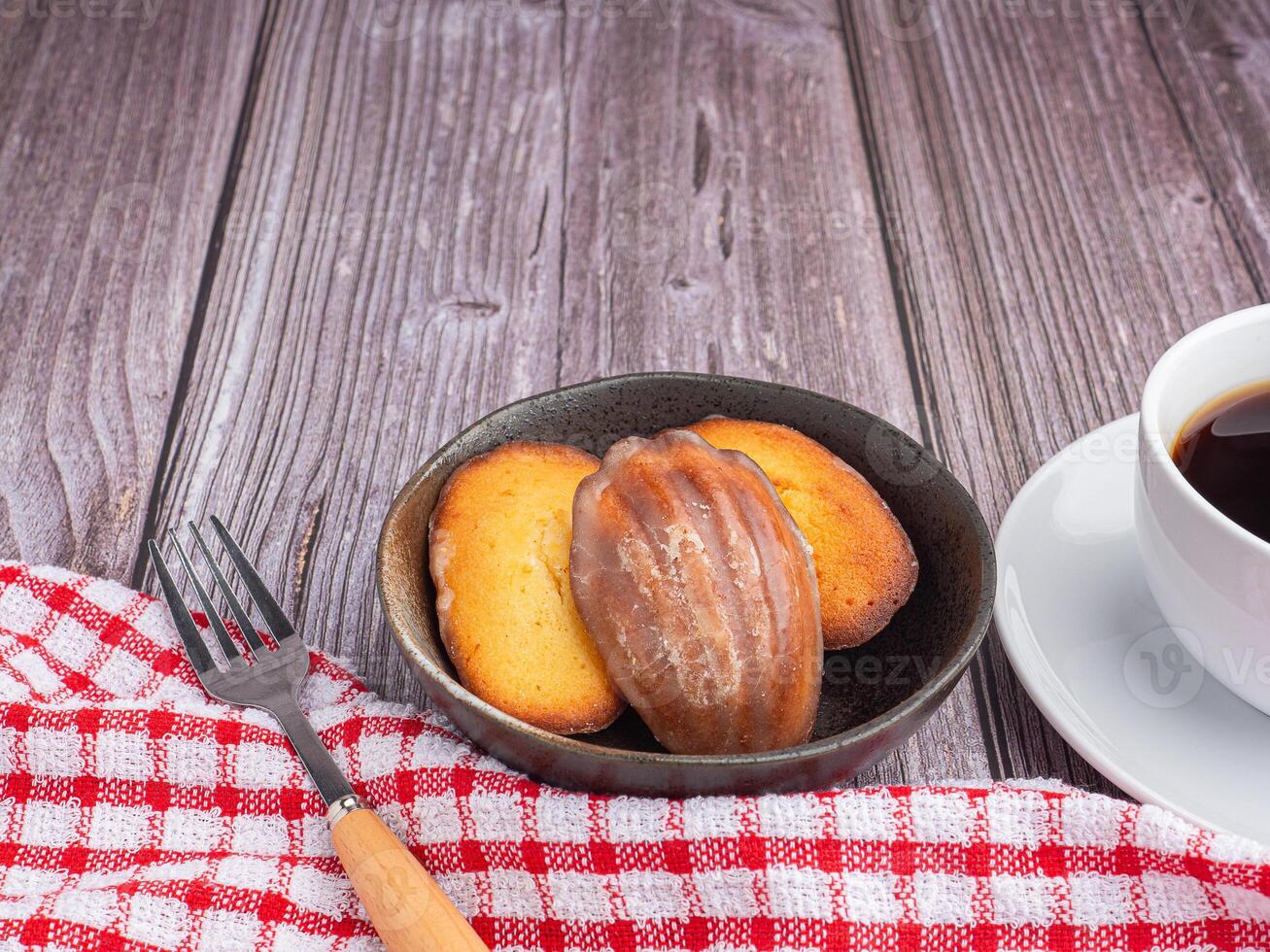 Lemon glaze madeleines in a bowl and a white coffee cup placed on a wooden table with copy space for text. Madeleine - homemade traditional French small cookie. Seashell sweet cakes photo