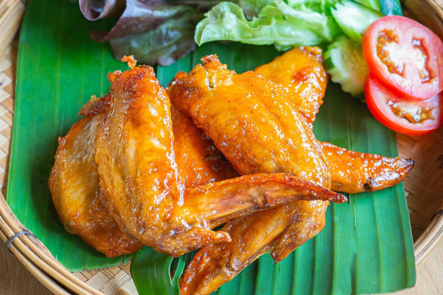 Chicken wing spicy, tomatoes, and lettuce on a banana leaf placed in a bamboo tray. Close-up photo