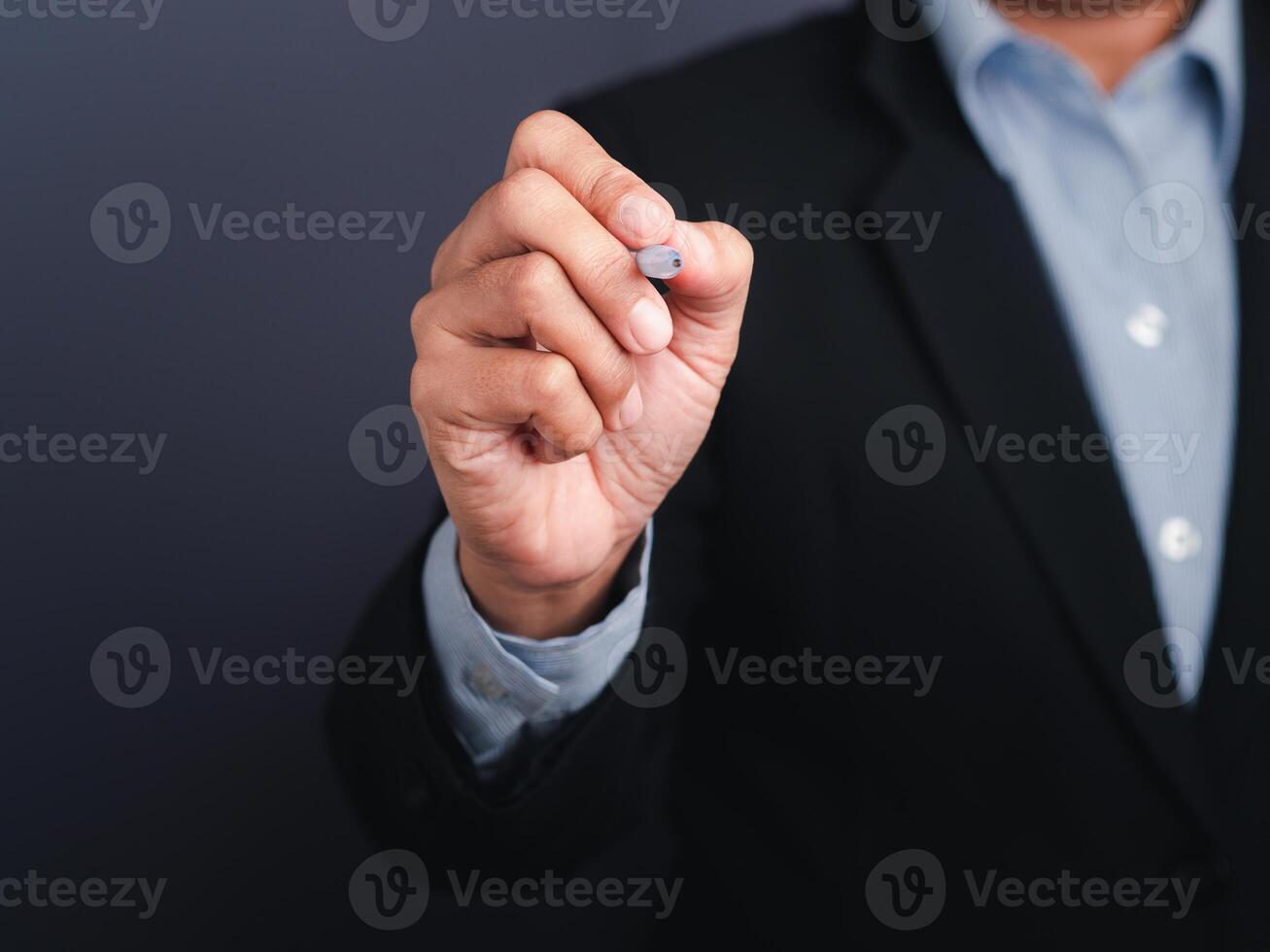 A businessman wearing a suit is holding a pen write or writing on the virtual screen or in the air against a gray background. Space for text. Close-up. Modern business background concept photo