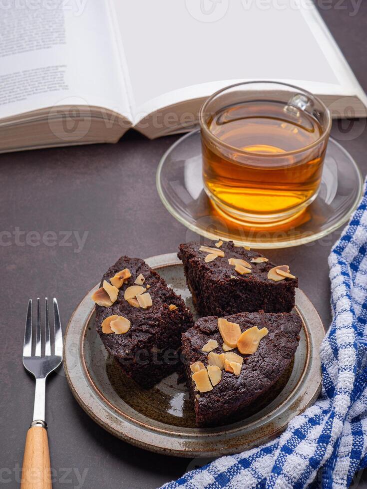 Pieces of fresh brownies on a plate with a book and teacup. Side view. Delicious chocolate pie. Dessert and relaxation concept photo