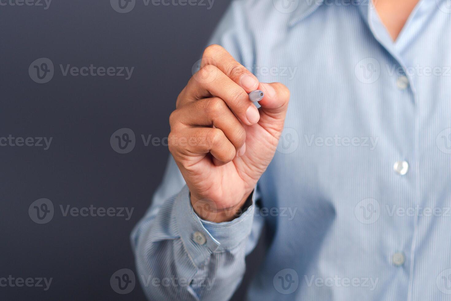 A businessman wearing a light blue shirt is holding a pen write or writing on the virtual screen or in the air against a gray background. Space for text. Close-up. Modern business background concept photo