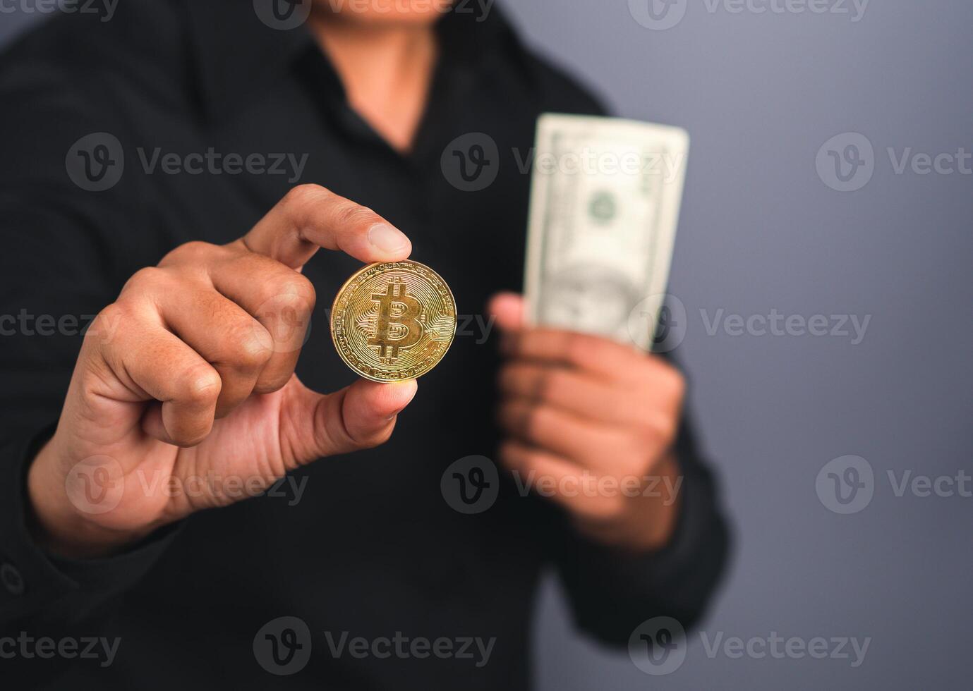 Portrait of an investor guy showing a bitcoin golden coin and US banknote with a gray background while standing in the studio. Space for text. Virtual cryptocurrency and future money concept photo