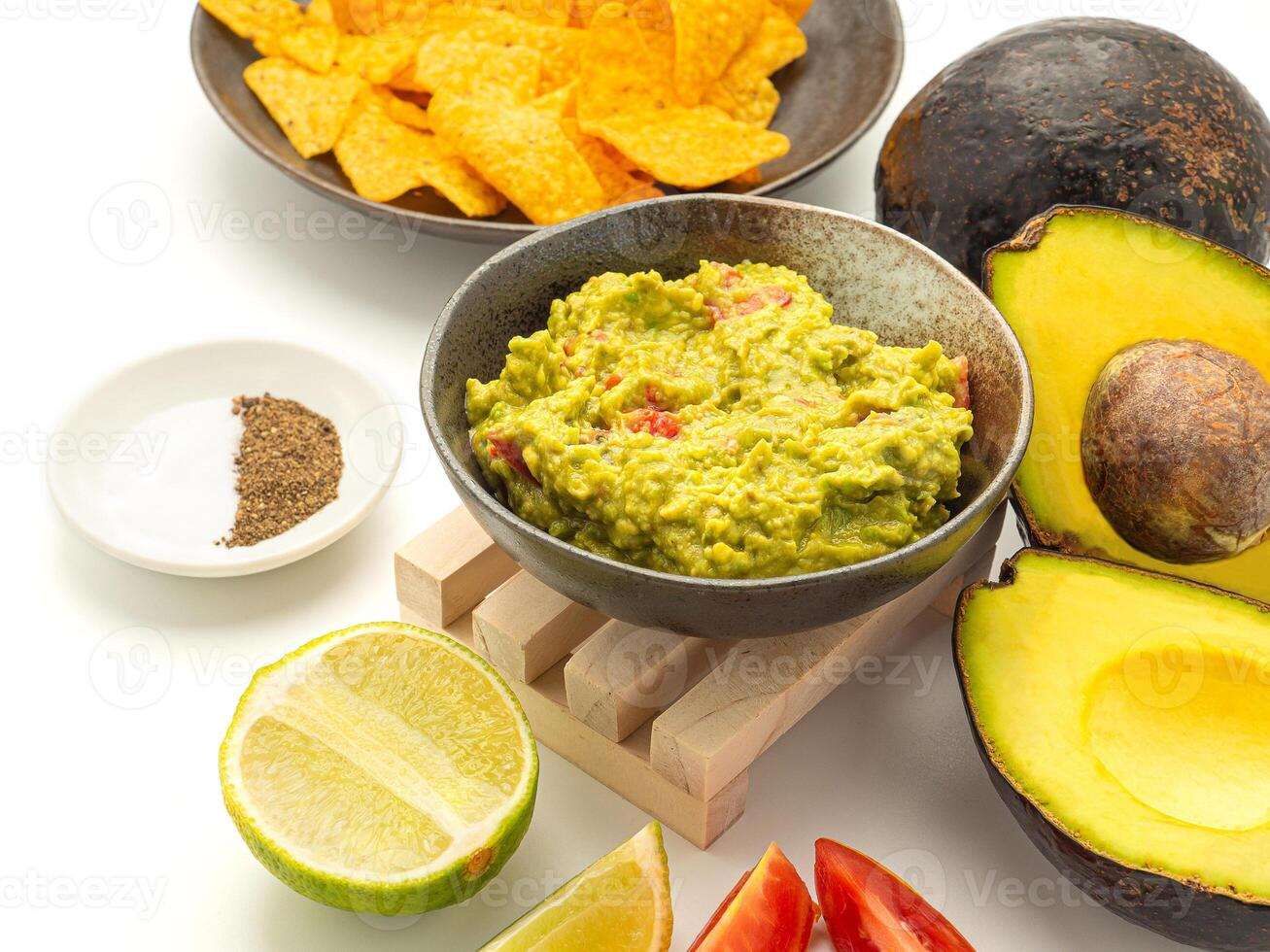 Fresh guacamole on a plate place with ingredients for homemade guacamole avocados, lemon, salt, pepper, and tomato on white background. Side view. Concept of traditional Mexican preparation photo