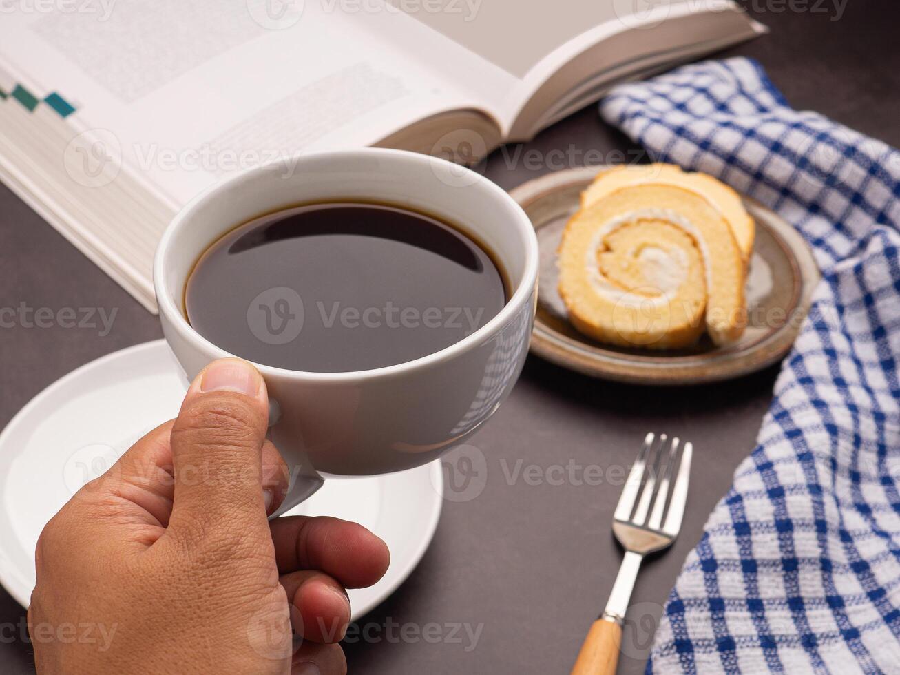 Hand holding a white coffee cup with a book, roll cakes on a plate and a cloth placed on a dark gray stone background. Close-up photo. Space for text. Concept of beverage and relaxation photo