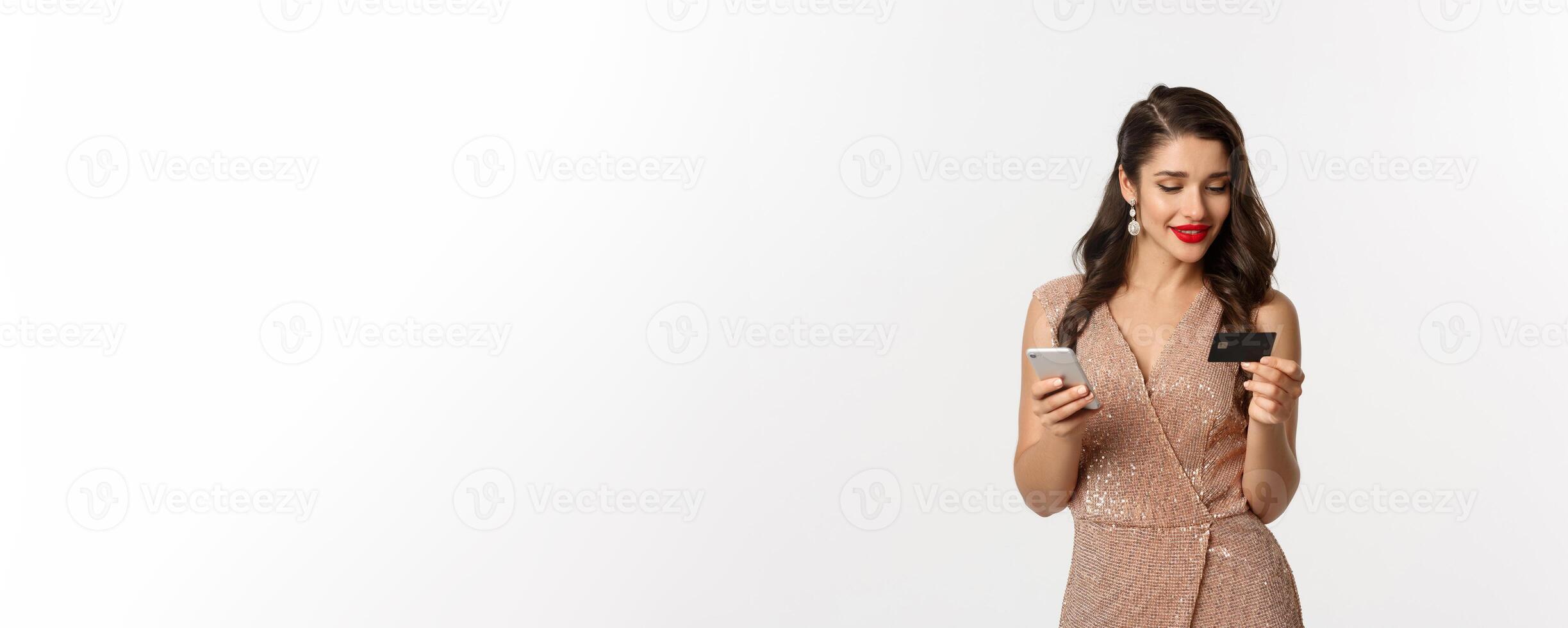 en línea compras y Navidad concepto. elegante joven mujer pago para Internet compra con teléfono inteligente y crédito tarjeta, en pie en lujo vestir terminado blanco antecedentes foto
