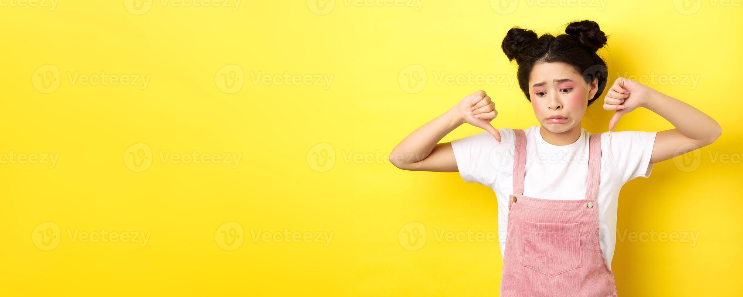 Asian beauty girl cringe from something disgusting, look away and showing thumbs-down to express dislike, negative emotions, standing in summer clothes on yellow background photo