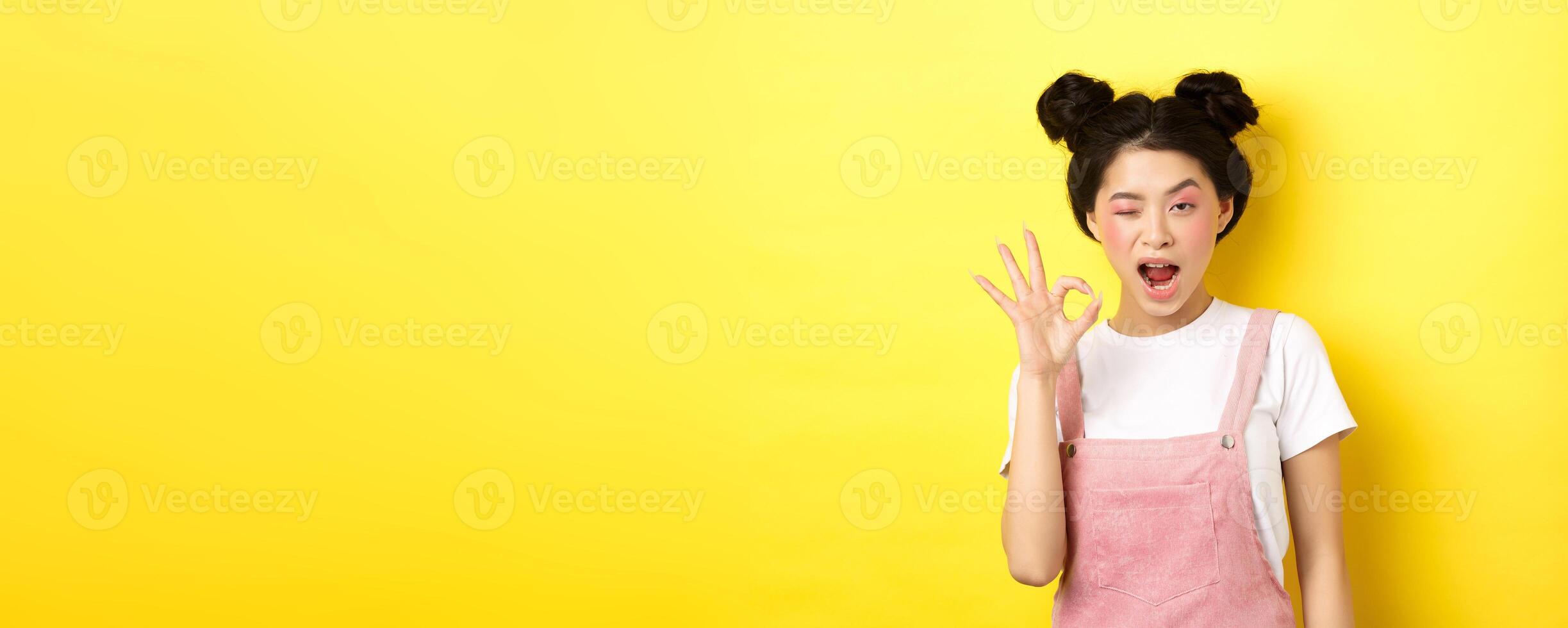 Alright. Confident asian female model with bright makeup and summer clothes, showing okay sign, winking and say yes, agree and approve, yellow background photo