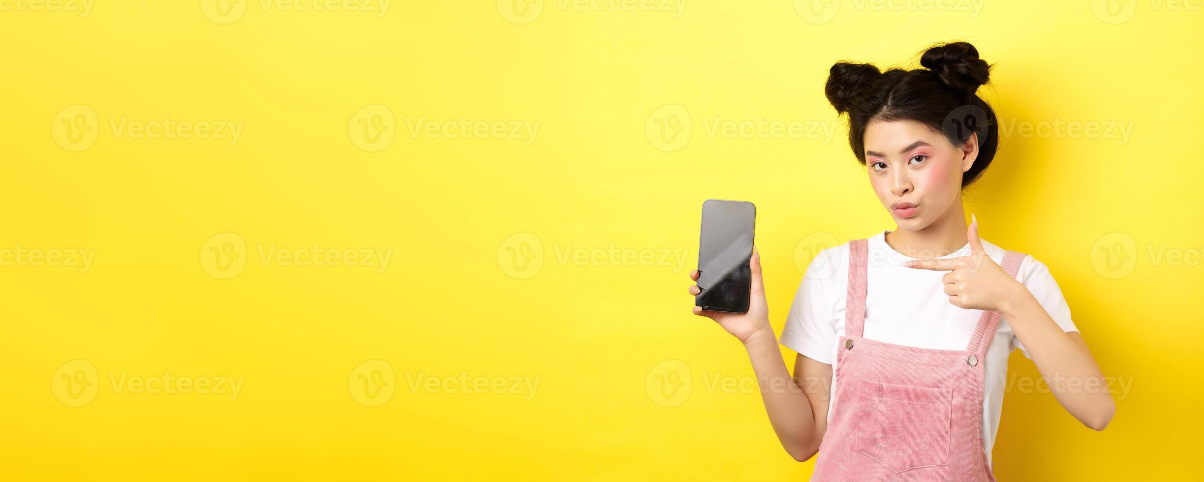 Online shopping concept. Stylish asian woman with pink makeup, pointing at empty smartphone sreen, showing good deal, yellow background photo