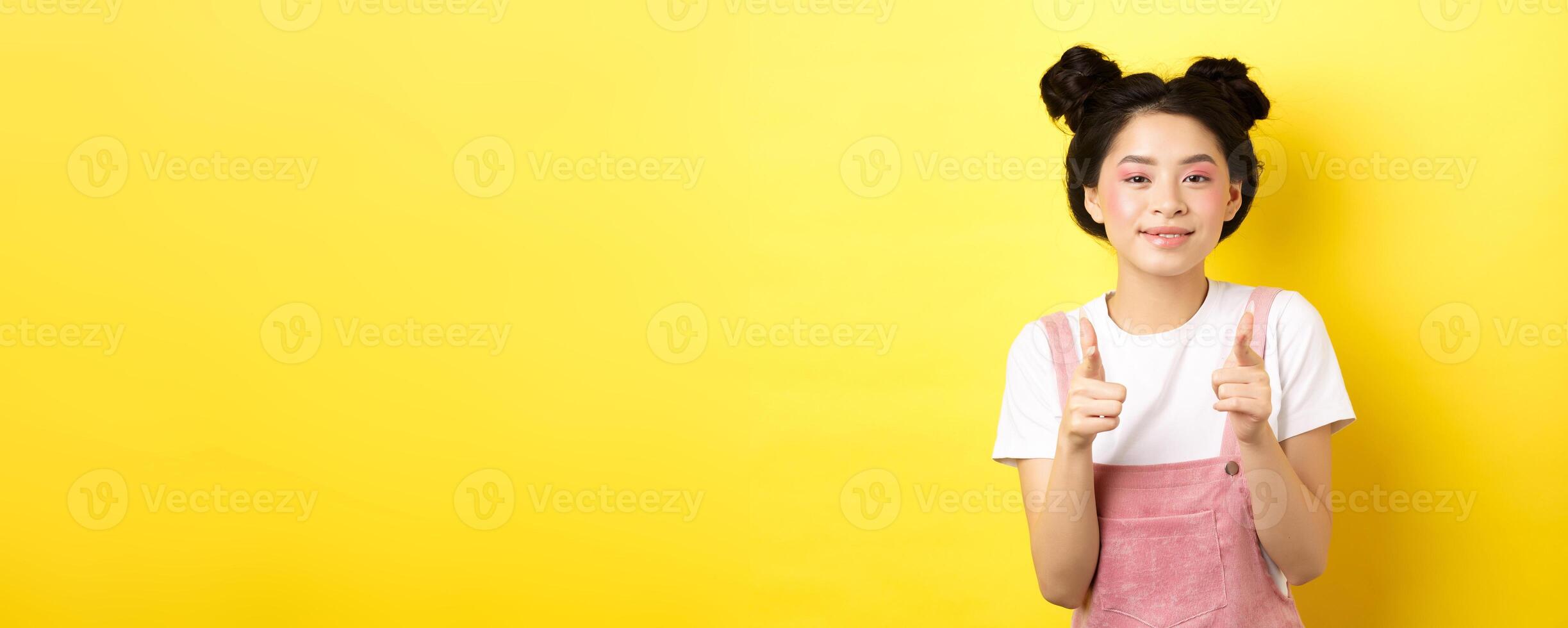 Cheerful teen asian girl pointing fingers at camera and smiling, encourage or invite you, making compliment, praise nice work, standing on yellow background photo