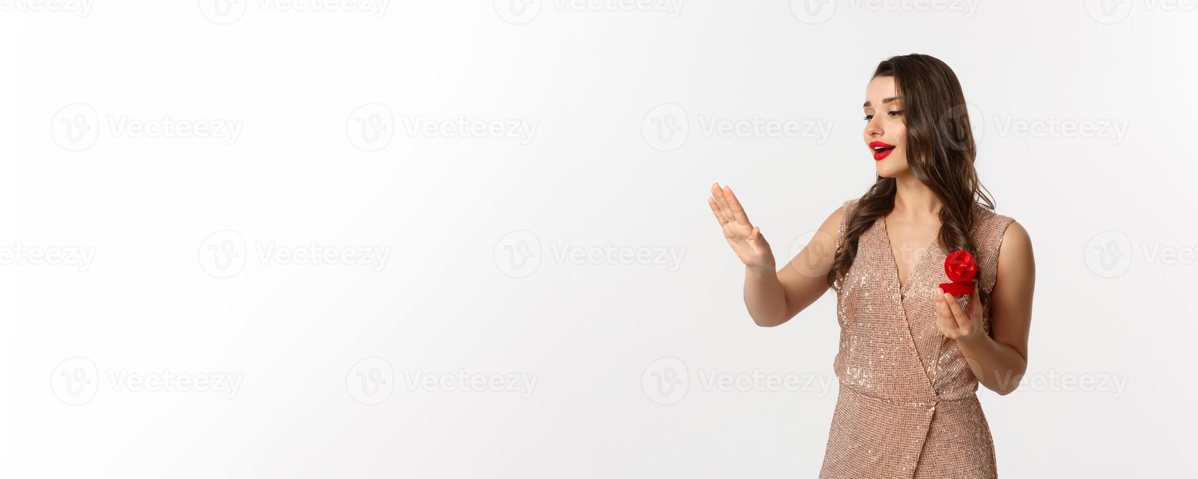 Beautiful woman in luxury dress, trying on engagement ring, saying yes to marriage proposal, looking dreamy at hand, standing over white background photo