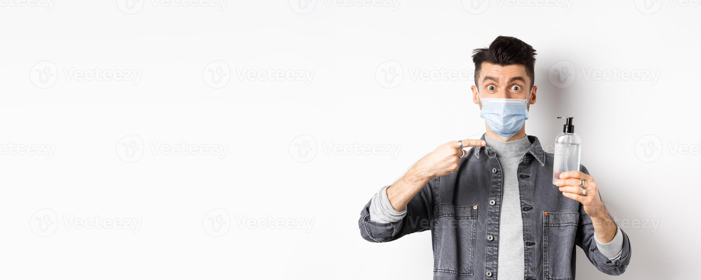 Health, covid and pandemic concept. Stylish guy in medical mask pointing at bottle with hand sanitizer, showing antiseptic, standing on white background photo