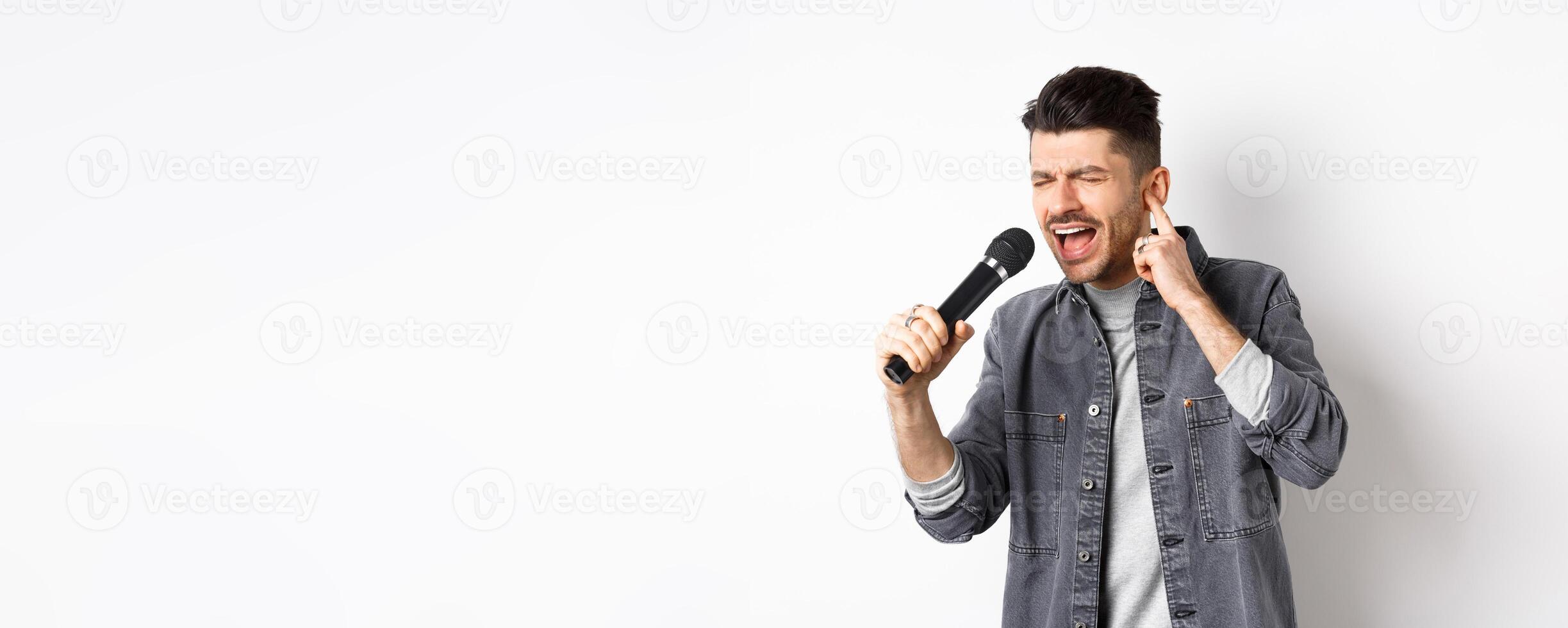 Handsome singer trying to reach high note, singing in microphone and shut one ear, standing on white background photo