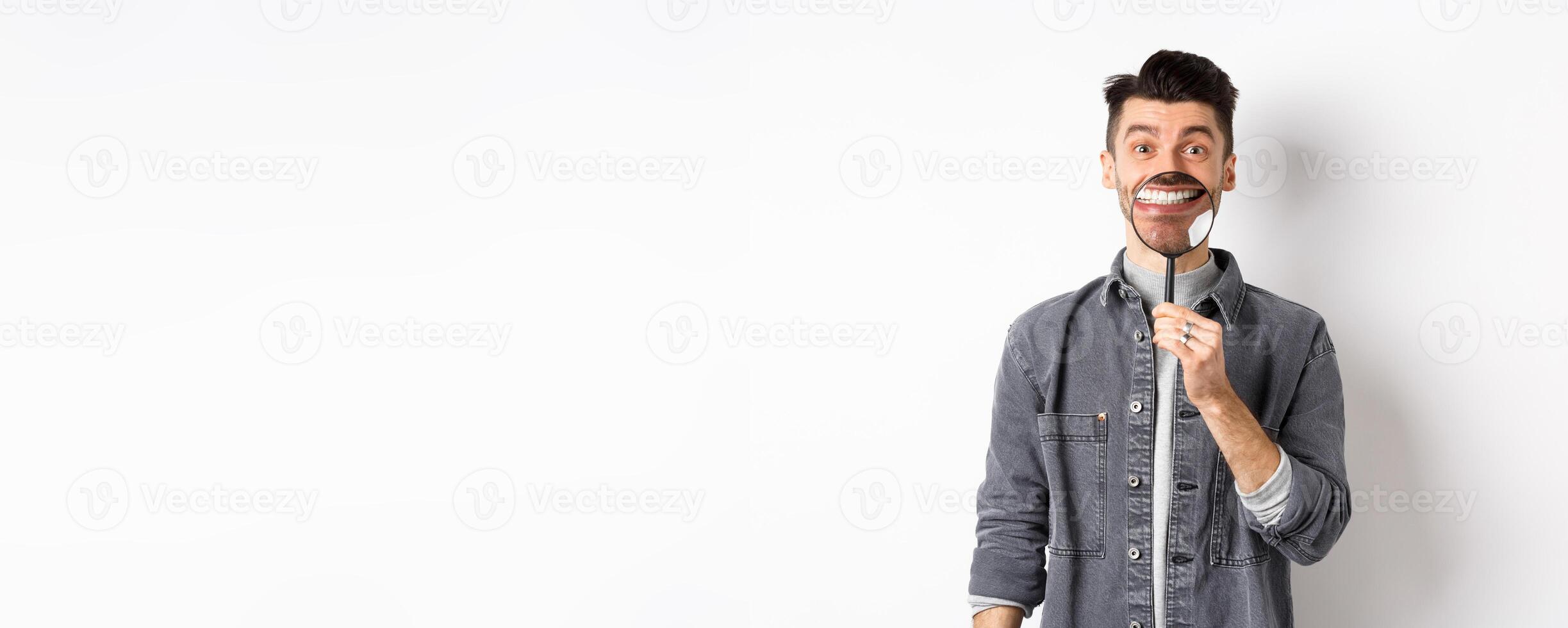 Handsome positive guy showing white perfect smile with magnifying glass, standing against white background photo