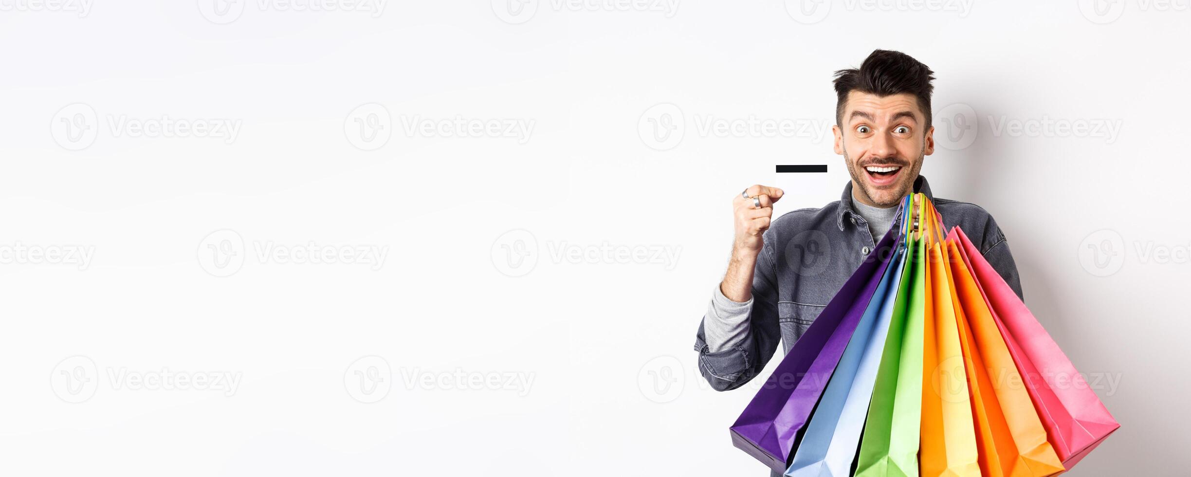 Happy guy shopaholic holding colorful shopping bags and showing plastic credit card, smiling excited, standing white background photo