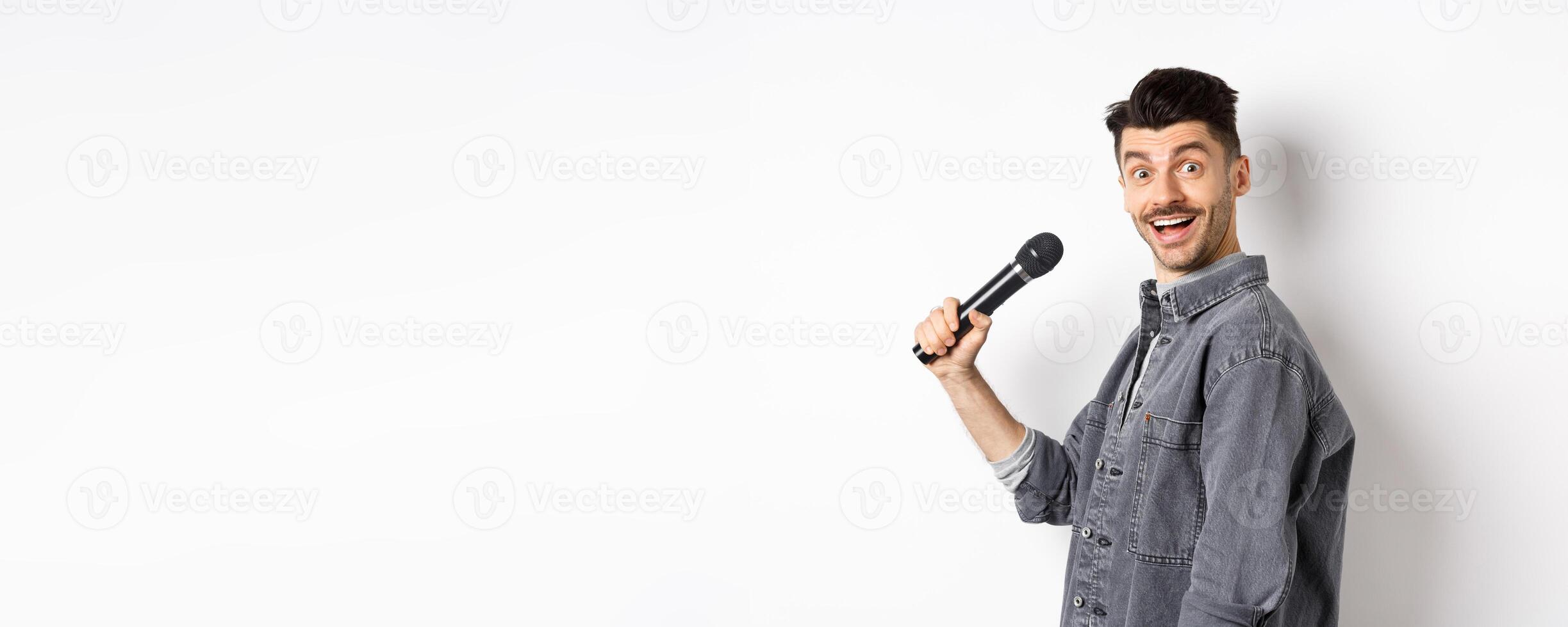 Profile of handsome smiling man holding mic, turn head at camera with excited face, singing karaoke and perform standup, standing on white background photo