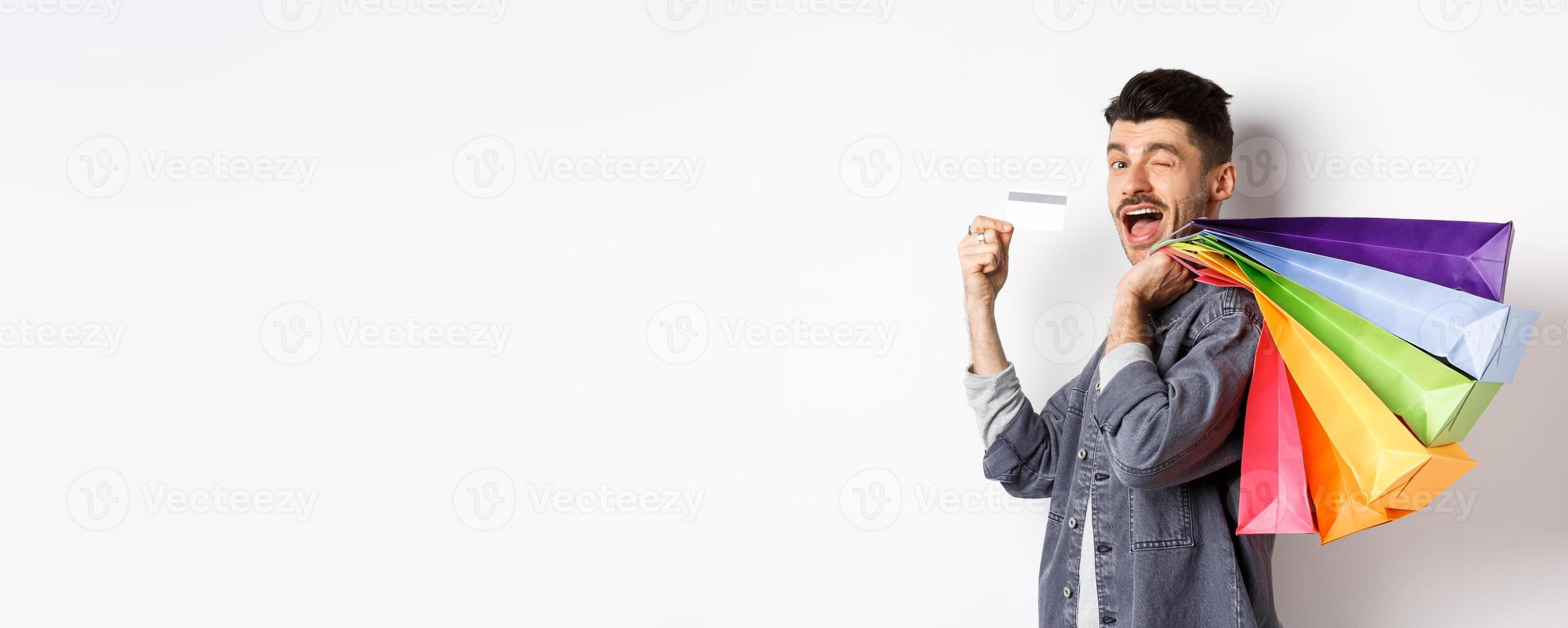 Happy shopper holding shopping bags over shoulder and showing plastic credit card, paying contactless, standing on white background photo