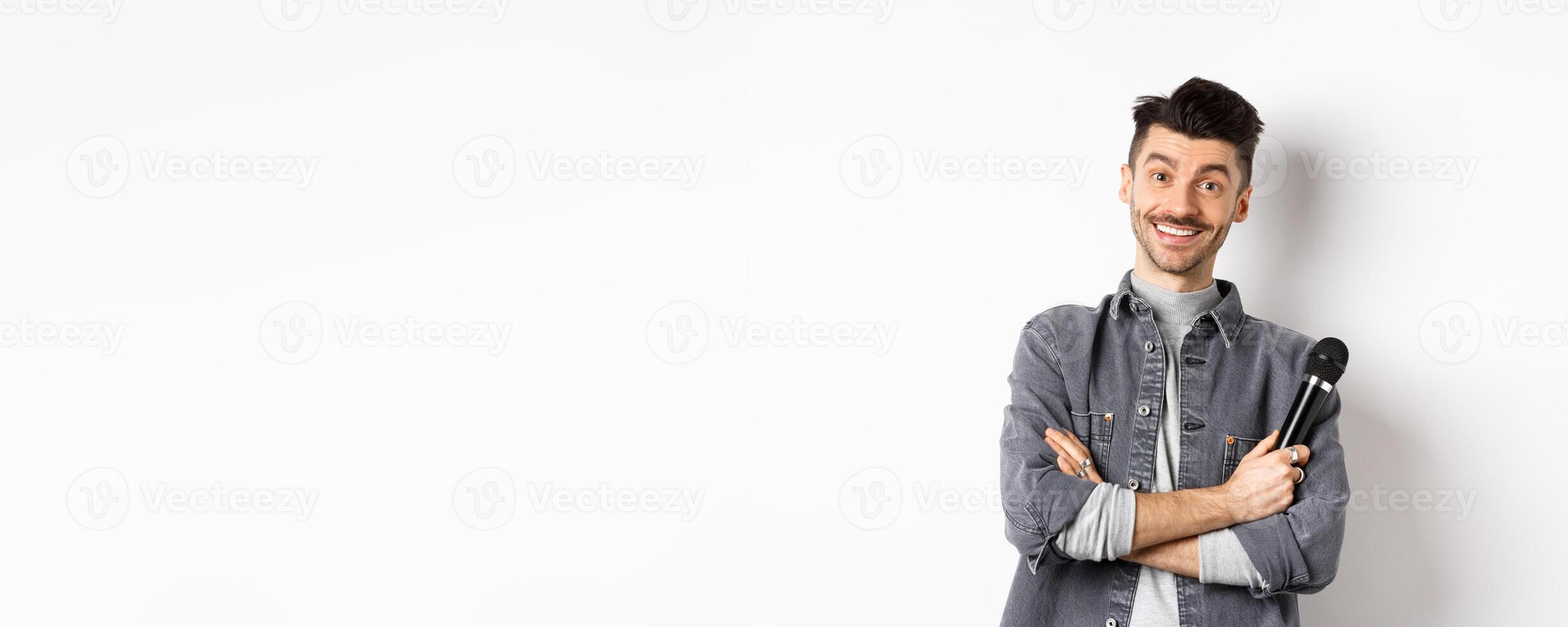 Handsome caucasian guy with moustache cross arms on chest, holding mic and smiling at camera, perform on stage with microphone, standing against white background photo